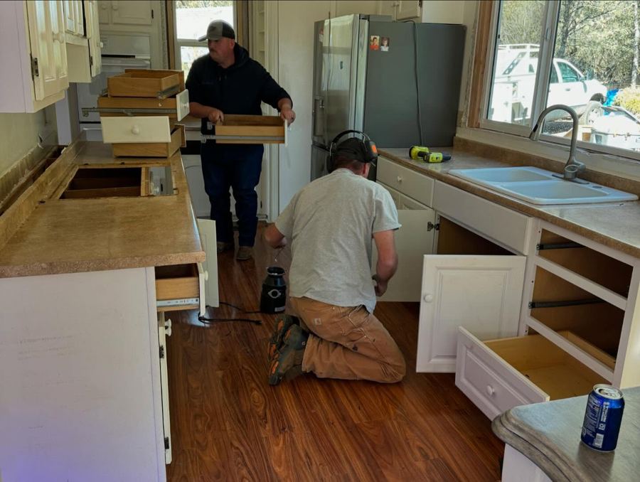 Kitchen demo: Chip in a grey tee and ear protection kneeling at the sink cabinet.
